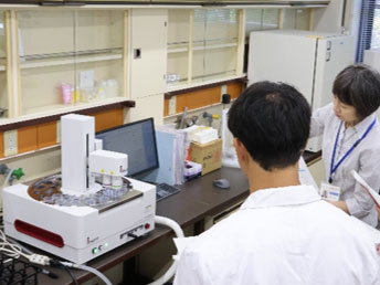 分析機器の前で研究所職員から説明を受ける学生たち